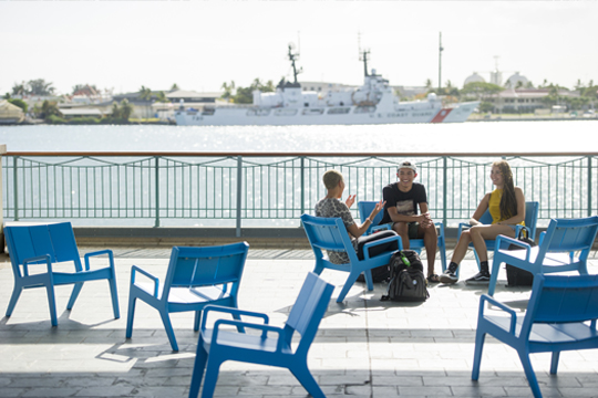 3 students chatting on blue chairs near the pier at Aloha Tower. Behind them, a boat is in the water.